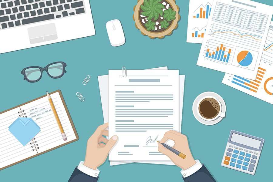 A man's hand signing a document with a cup of tea, calculator, an eyeglass, laptop, and business reports around him.