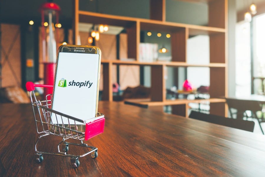 Shopifyapp on Mobile Phone screen and in a shopping cart on wooden table.