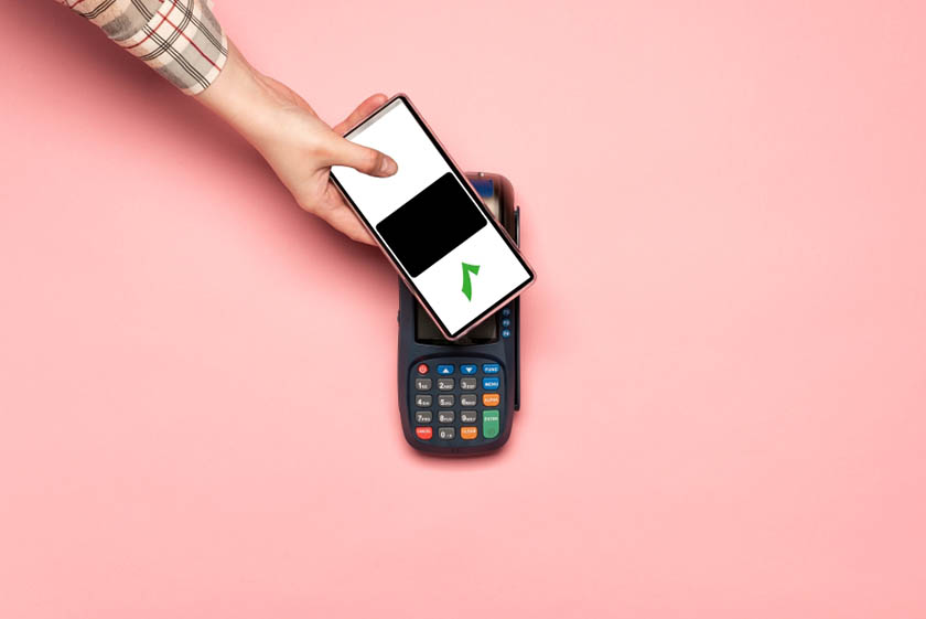 A smartphone being held up to a contactless payment terminal on a pink background.