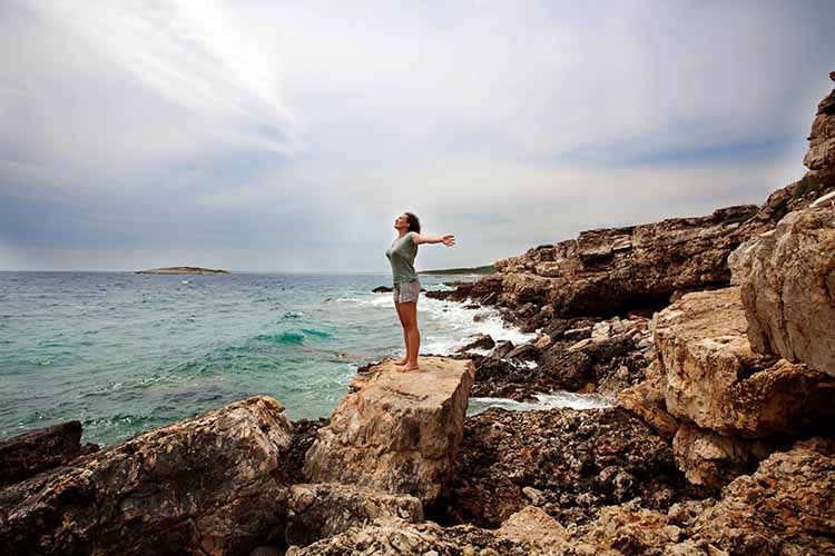 一个穿着短裤和t恤的赤脚女人站在多岩石的海岸线上，海浪拍打着岩石，她举起双手，呼吸着咸咸的海洋空气。