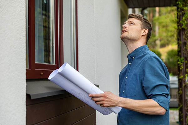 一位房地产估价师站在一户人家外面，手里拿着一卷纸，抬头望向屋顶。
