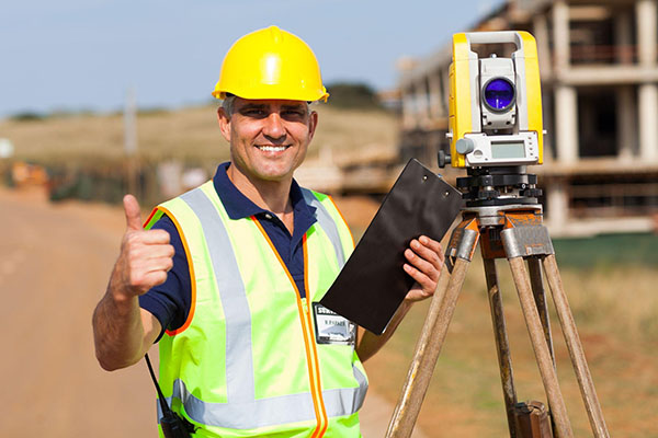 土地测量师头戴黄色安全帽，身穿安全背心，站在测量设备旁竖起大拇指。